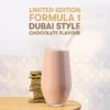 Promotional image showing a tall glass of pink chocolate shake with a curved straw, next to a stack of pistachio cookies on a wooden table, with gold text reading 'LIMITED EDITION FORMULA 1 DUBAI STYLE CHOCOLATE FLAVOUR' above.