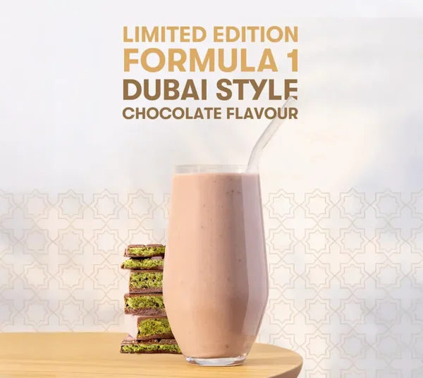 Promotional image showing a tall glass of pink chocolate shake with a curved straw, next to a stack of pistachio cookies on a wooden table, with gold text reading 'LIMITED EDITION FORMULA 1 DUBAI STYLE CHOCOLATE FLAVOUR' above.